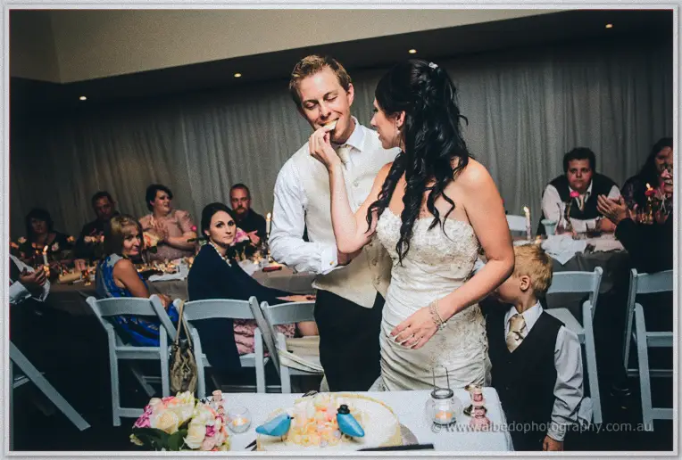 Sarah feeds Daniel a taste of cake while seated guests laugh under warm reception lights.