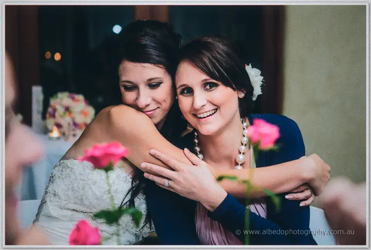 Sarah hugs a laughing bridesmaid in pearls, pink roses soft in the foreground at Brookleigh.