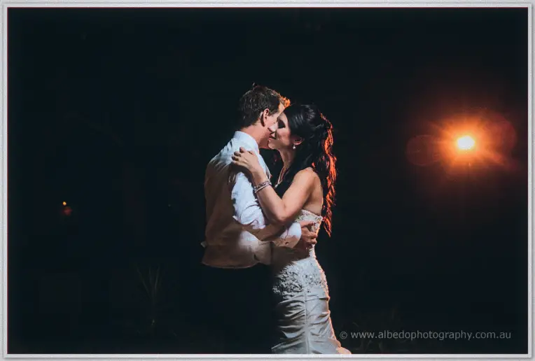 Sarah kisses Daniel mid-dance, a lone lamp flaring warm orange in the darkness behind them.