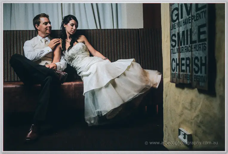 Weary at day's end, Sarah slumps against Daniel on a striped lounge beside a painted sign.