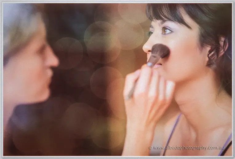 Bride Sarah having powder swept across her cheek with a brush, her reflection soft in the mirror