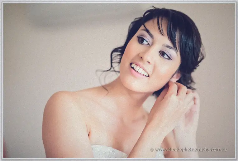 Bride Sarah smiling as she fastens a crystal earring, her strapless lace gown softly lit