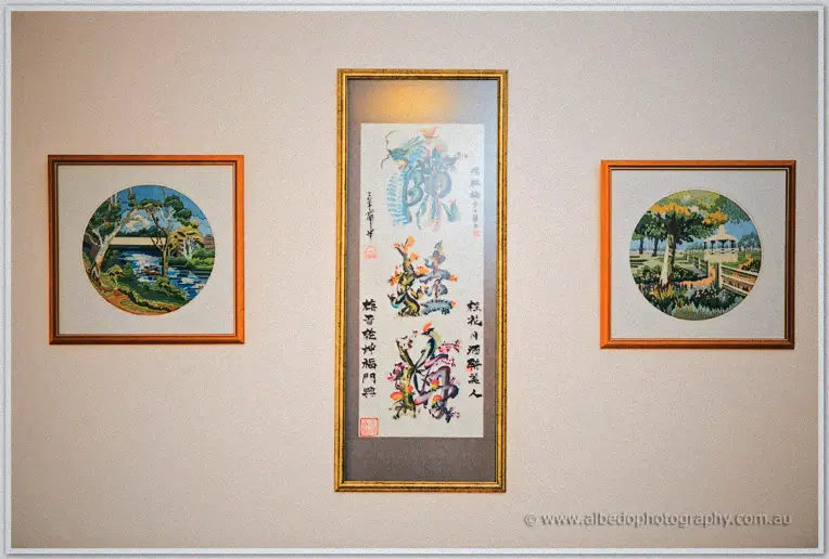 Framed needlework scenes and a gold-framed calligraphy scroll hanging on a cream venue wall