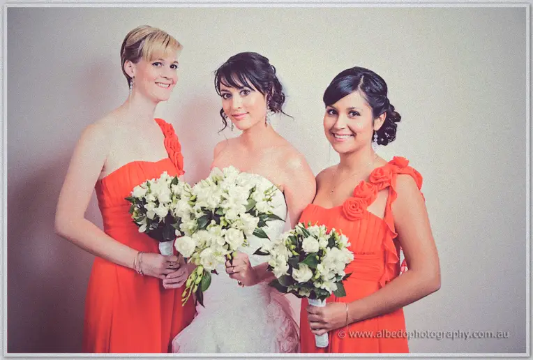 Bride Sarah with her two bridesmaids in orange dresses holding white bouquets against a pale wall
