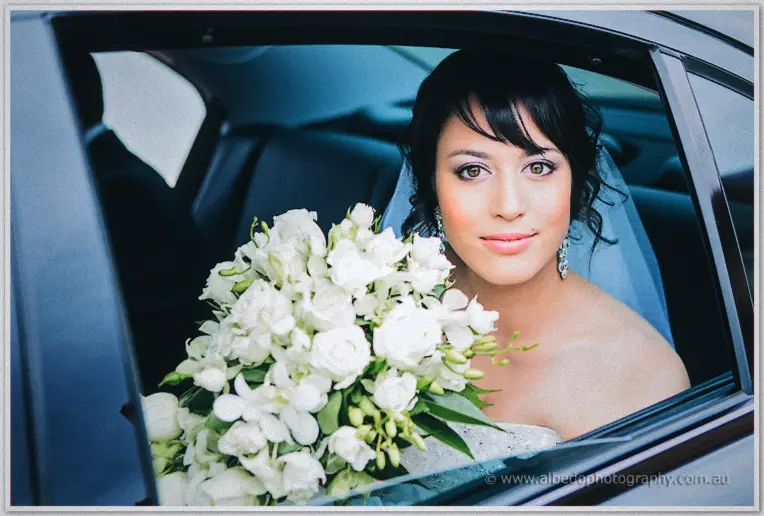 Bride Sarah peering through the car window, her white rose and orchid bouquet filling the frame