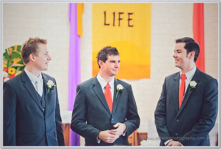 Groom Shannon and groomsmen in charcoal suits with coral ties exchanging grins beneath the banner