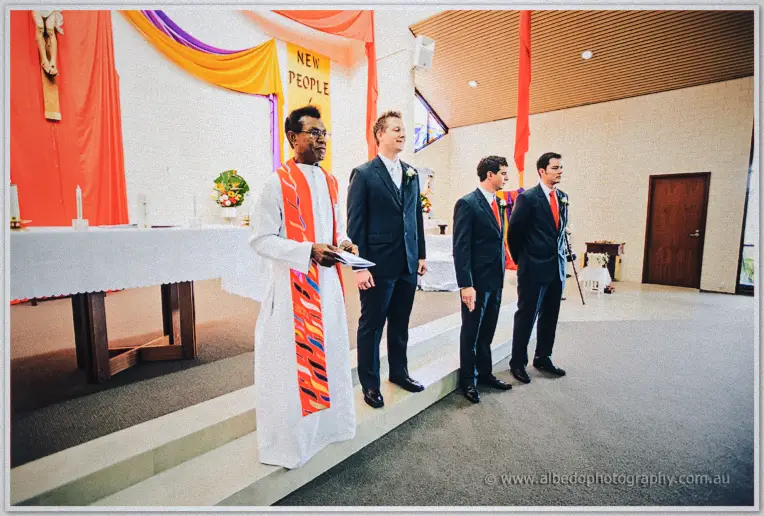 Groom Shannon beside the robed priest with groomsmen behind him before the cloth-draped altar