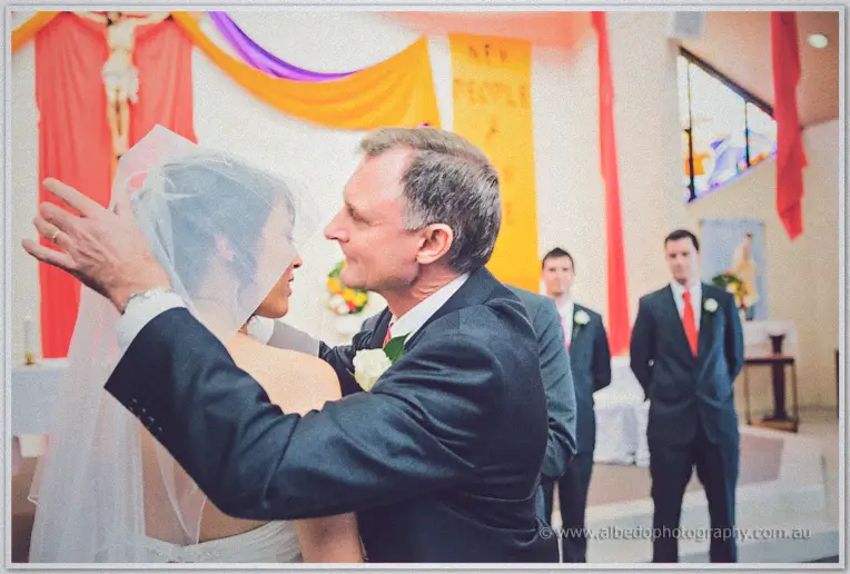 Sarah's father embracing her beneath the veil at the altar as the groom and groomsmen watch