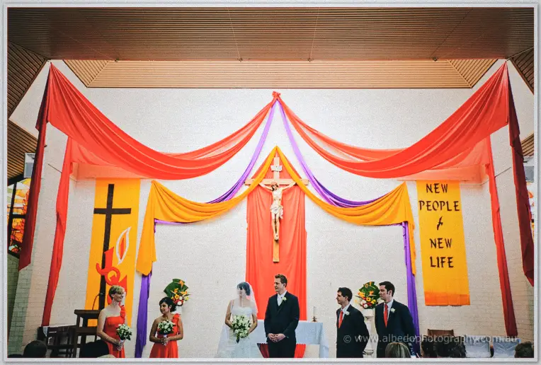 Wide altar scene of Sarah and Shannon before the priest beneath the crucifix and colorful drapes