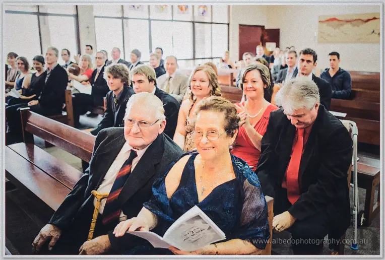 Congregation smiling from the wooden pews, an elderly couple in the front row holding the booklet