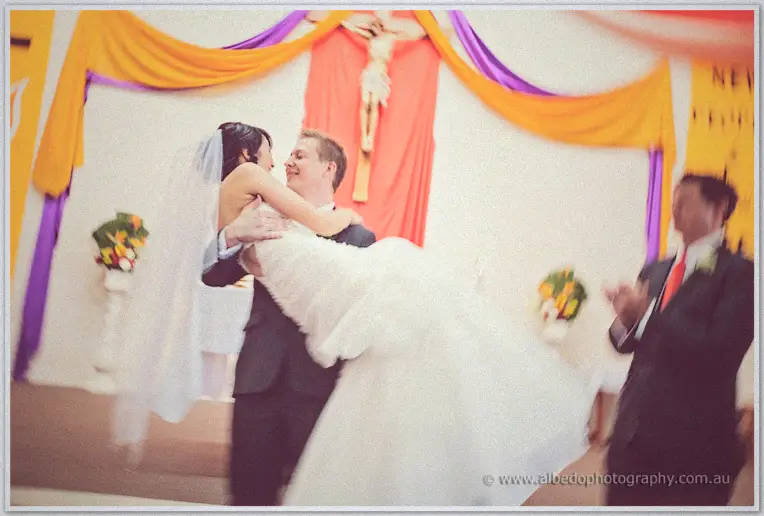 Shannon sweeping Sarah up into his arms on the altar as a groomsman applauds, both laughing