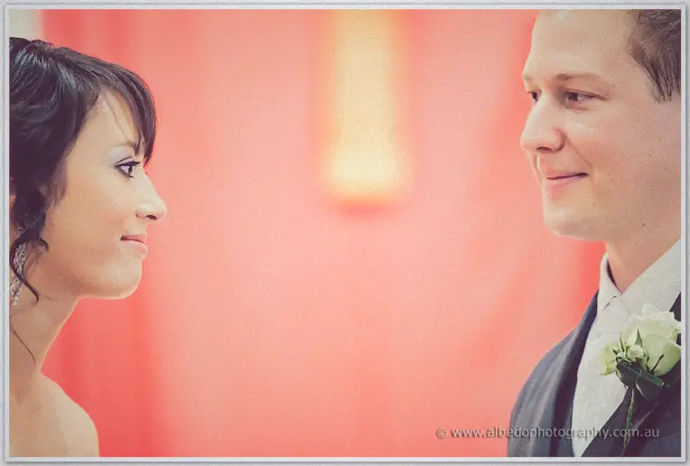 Profile of bride and groom facing each other before the red-draped altar as Sarah smiles up