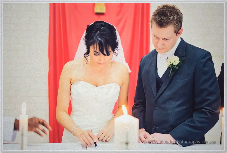 Bride Sarah signing the marriage register with Shannon beside her, a lit candle glowing nearby