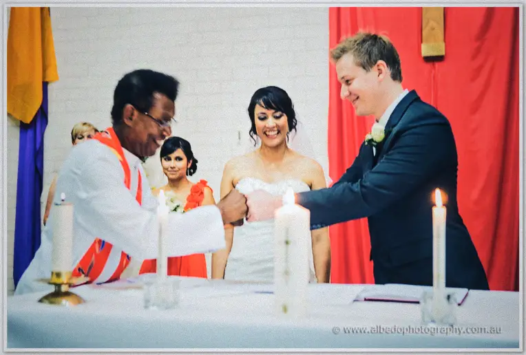 Priest shakes Shannon's hand at the altar as Sarah smiles between them, candles and red drapes aglow