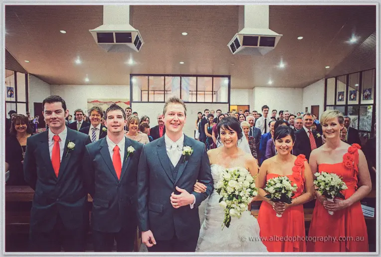 Bridal party in coral gowns and red ties lines the church aisle behind Sarah and Shannon, guests standing