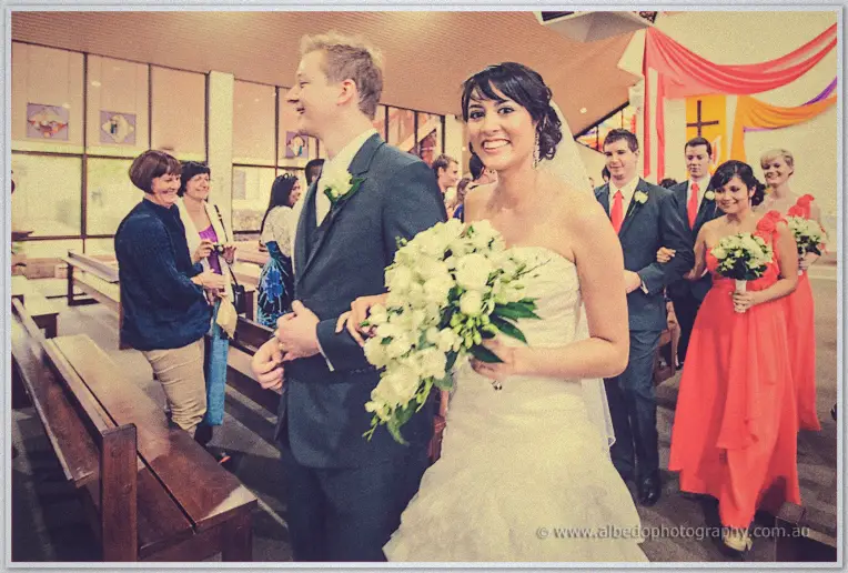 Sarah beams carrying white roses as she and Shannon walk back up the aisle past smiling guests