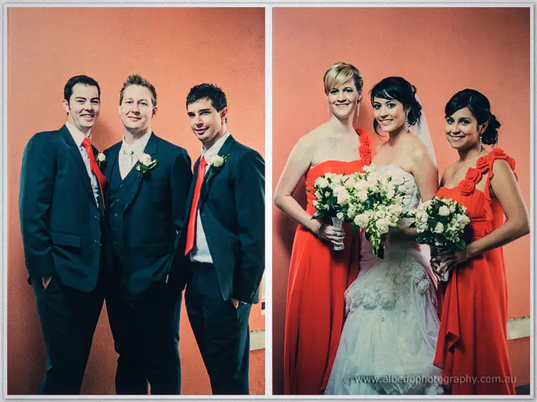 Groomsmen in red ties and bridesmaids in red gowns flank the newlyweds for portraits on the coral wall