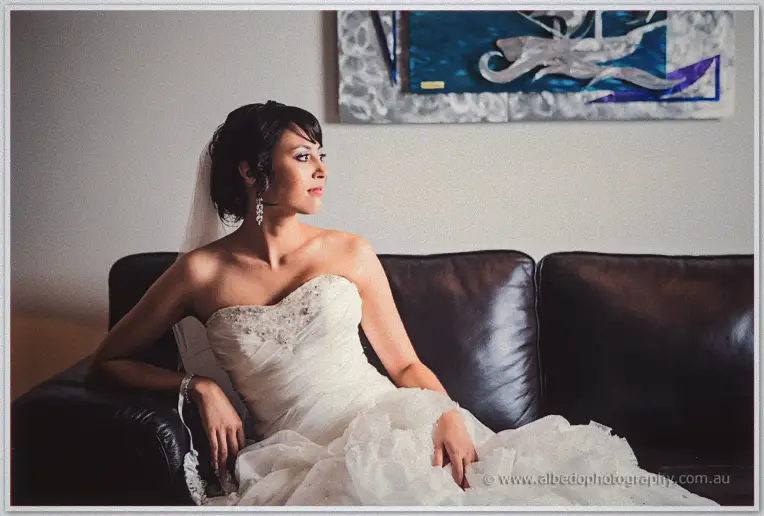 Sarah pensive on a dark leather lounge in her beaded gown, veil trailing, abstract art behind