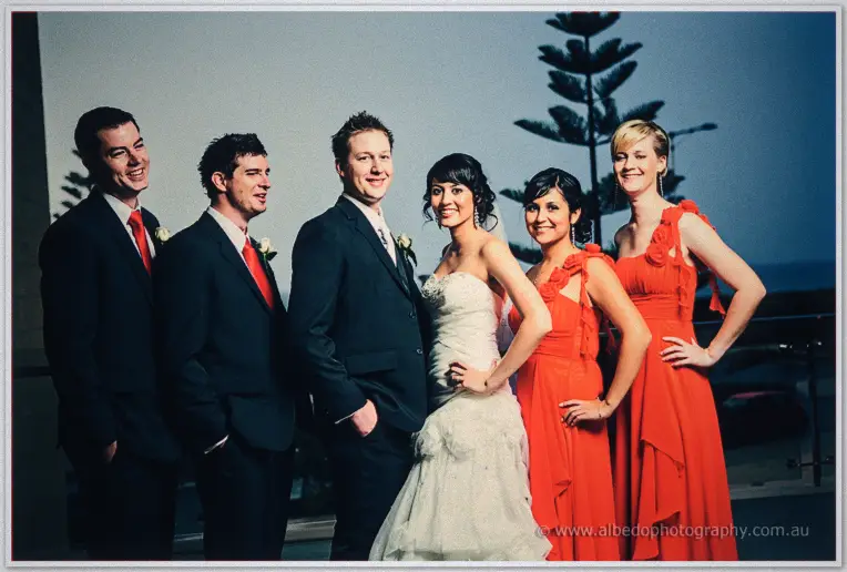 Bridal party in red gowns and ties poses at dusk beside a Norfolk pine, hands on hips and grinning