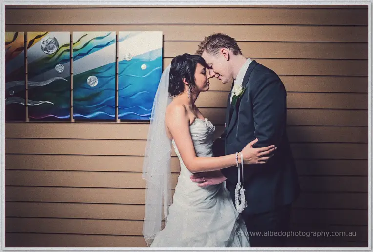 Sarah and Shannon nuzzle foreheads before a timber wall of ocean-toned panels, veil drifting