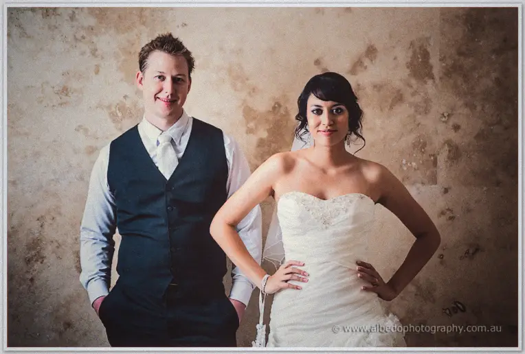 Shannon hands in pockets and Sarah hands on hips smile at the camera before a weathered plaster wall
