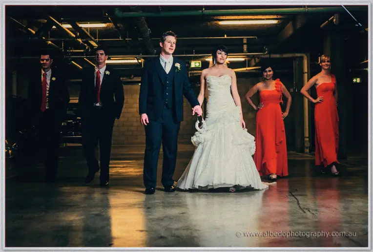 Bridal party strides moody underground carpark rows, red gowns and ties glowing under strip lights