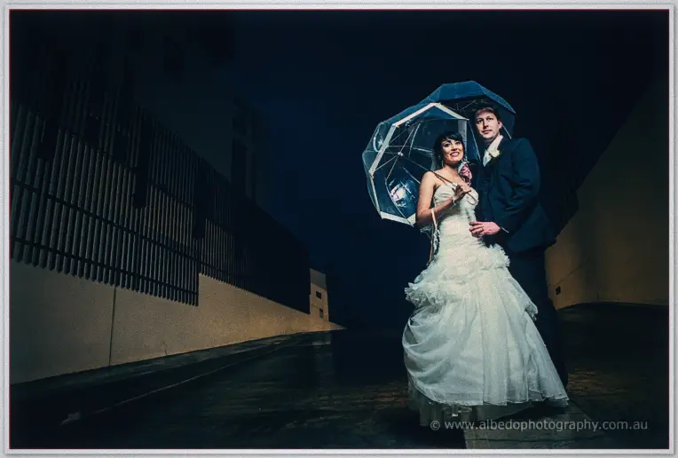 Bride and groom sheltering beneath a clear umbrella on a dark wet street at night