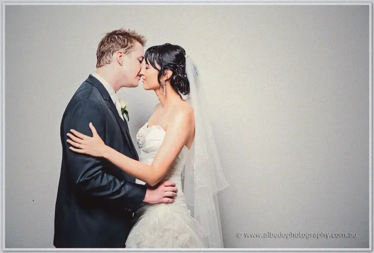 The newlyweds share a gentle kiss against a plain grey backdrop, veil spilling over her shoulder