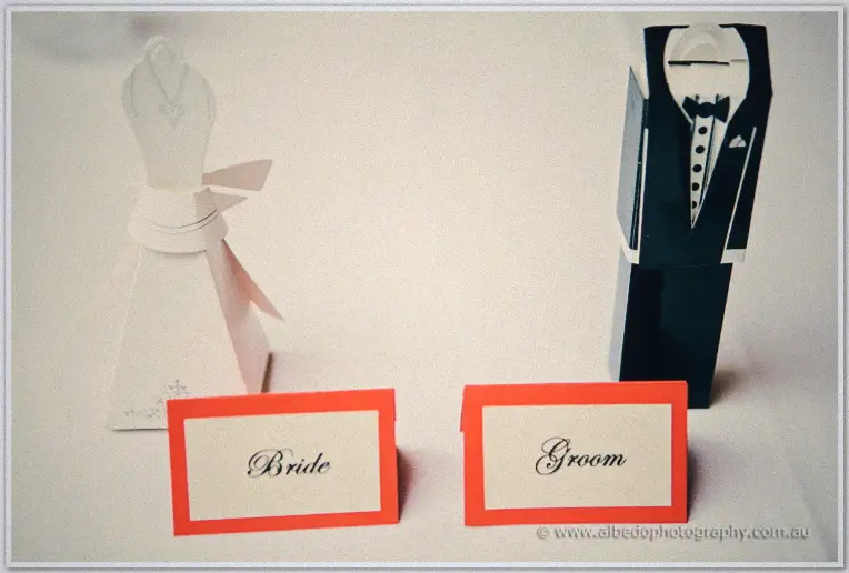 Bride and groom dress-shaped boxes sit behind red-framed Bride and Groom place cards on white linen