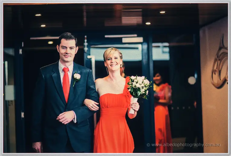 A groomsman and a blonde bridesmaid in red walk arm in arm into the reception, grinning