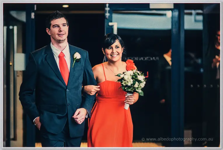 Another groomsman escorts a smiling dark-haired bridesmaid with a rose bouquet through glass doors