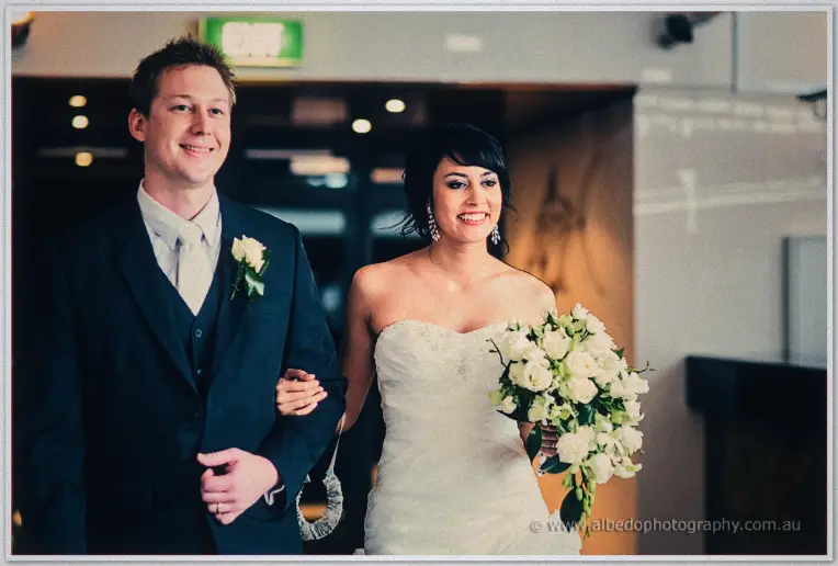 Sarah on Shannon's arm enters the reception beaming, white rose bouquet in hand