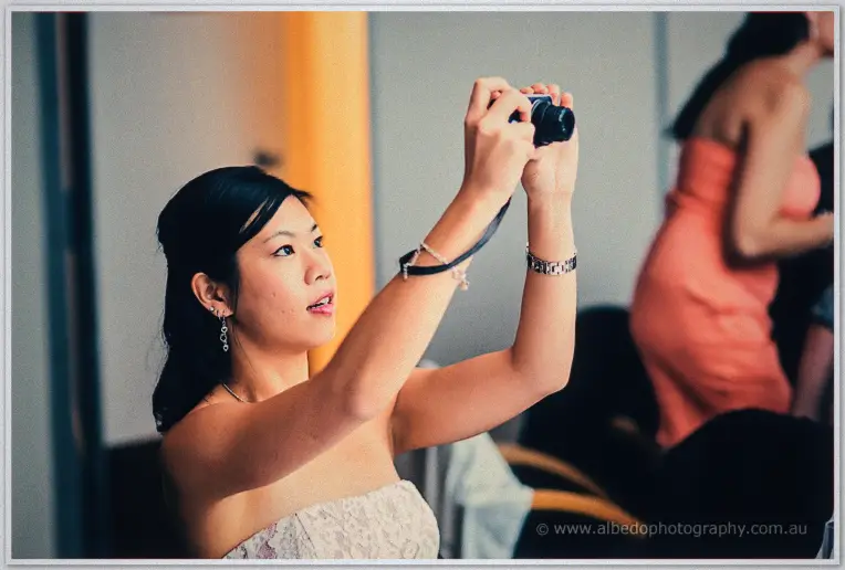 A guest in a strapless dress raises a compact camera to snap the celebrations, reception bustle behind