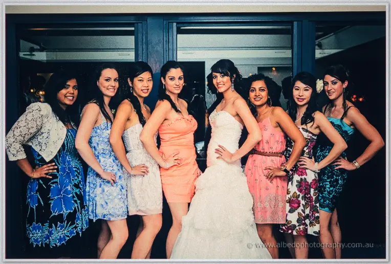 Sarah and seven friends in bright cocktail dresses pose hands-on-hips before glass doors at night
