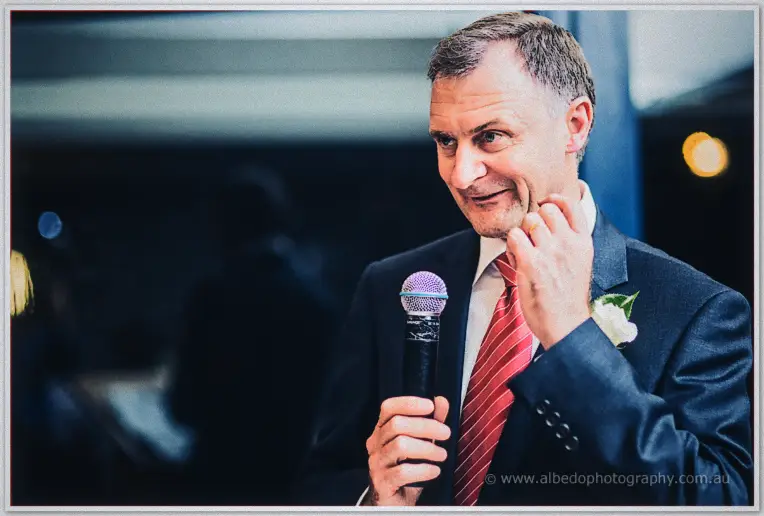 An older groomsman in a red-striped tie and white rose boutonniere touches his ear mid-speech