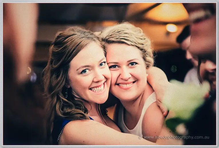 Two smiling friends hug cheeks at the reception, one in blue and one in cream under warm lights