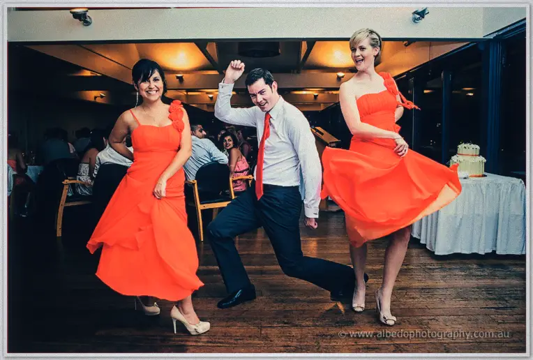 A groomsman in a red tie cuts a dance move between two bridesmaids twirling in scarlet gowns