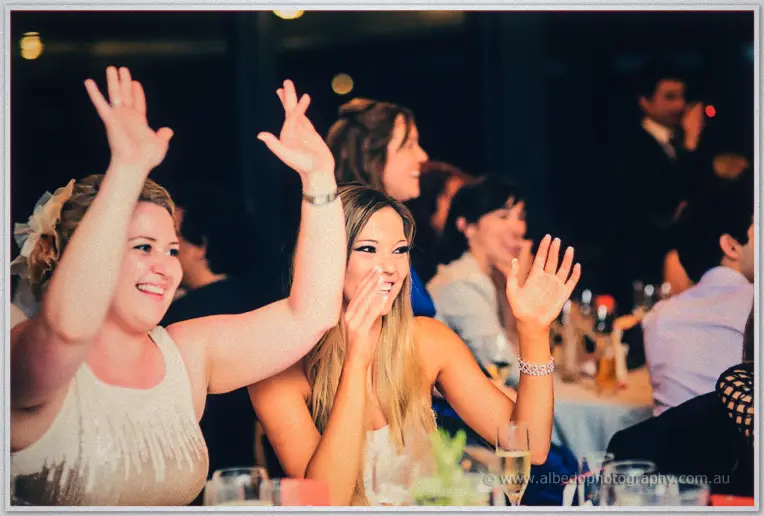 Guests wave and clap from their tables, champagne flutes raised, one covering a delighted grin