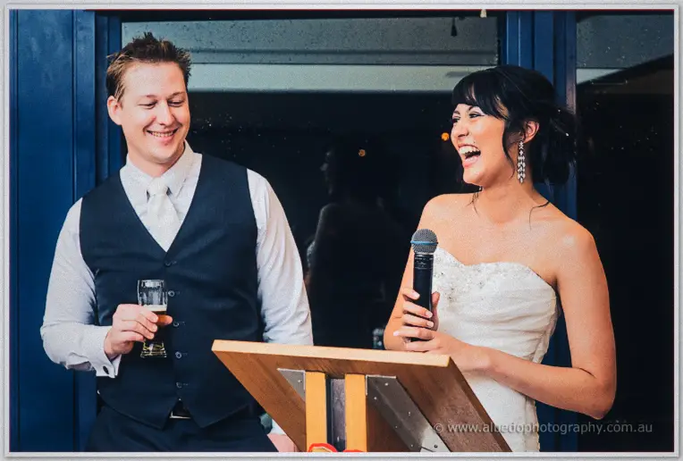 Sarah laughs into the mic at the lectern while Shannon watches with a beer beside blue shutters