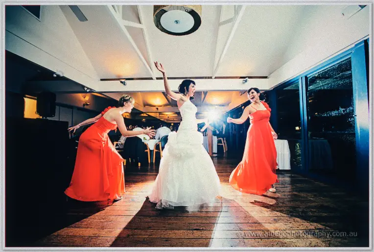 Bridesmaids in coral swing Sarah's skirt as she spins, guests watching under white rafters