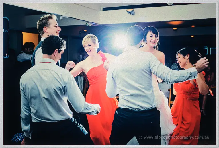 Guests in coral and white shirts dance in a circle around Sarah, flare blazing overhead
