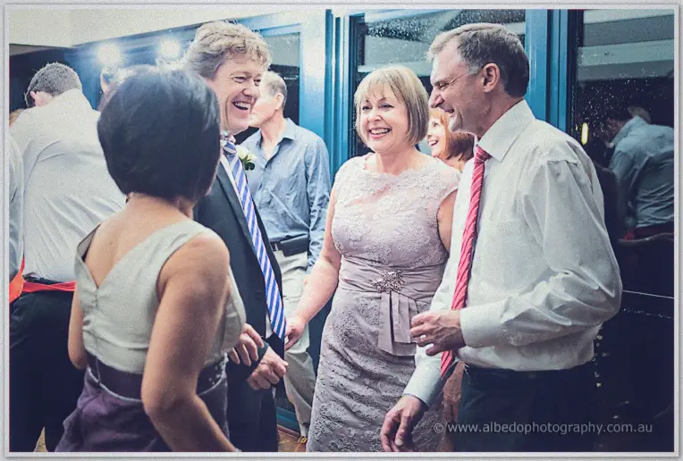 Older guests in lace and striped ties laugh together on the dance floor by blue-framed doors