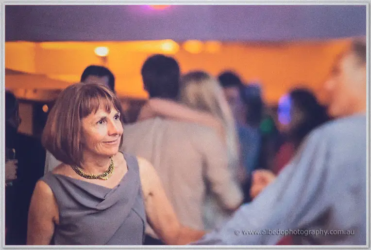 A guest in grey with a beaded necklace smiles amid dancing, warm lights blurring behind