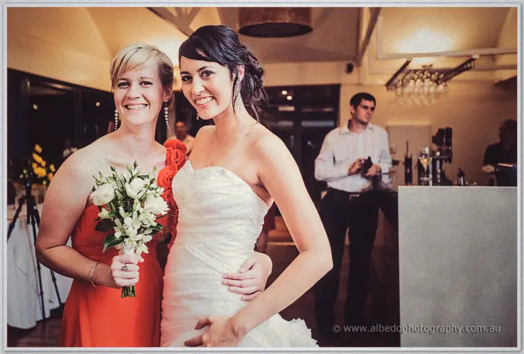 Bride in white and a bridesmaid in coral laugh arm in arm under warm lights at the Red Herring reception