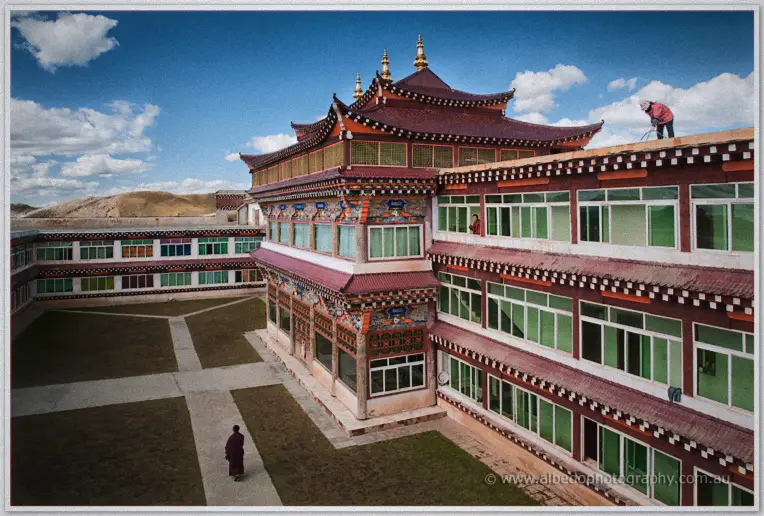 Craftsman repairs the ornate roof of a Tibetan monastery courtyard as a monk crosses below