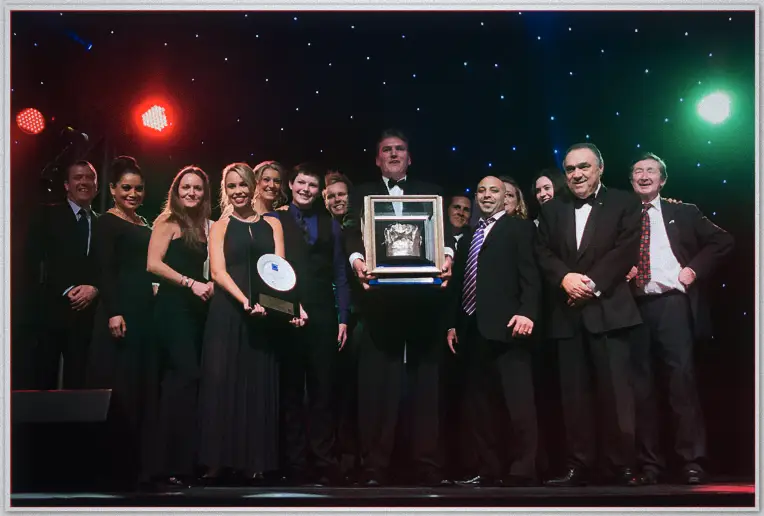 Award winners in evening wear crowd the lit stage as one man holds up a cased trophy
