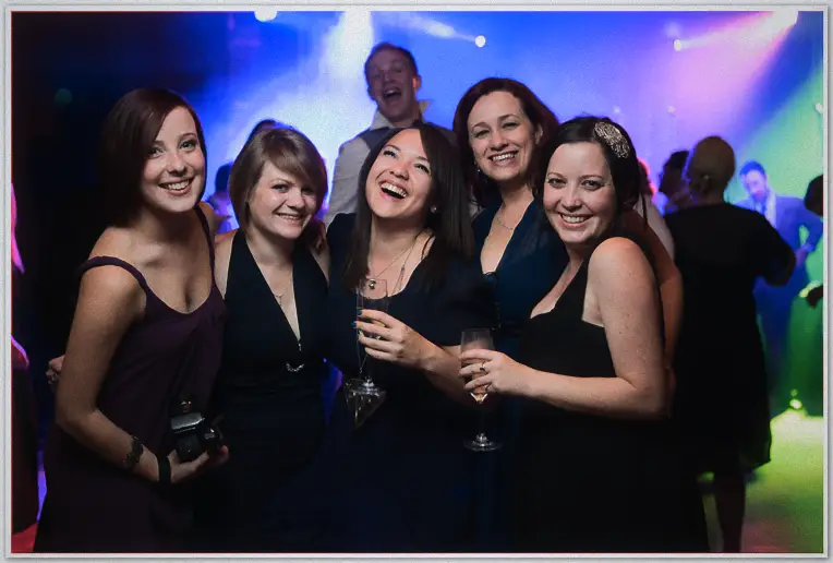Five women in black cocktail dresses laugh together, champagne glasses raised on the dance floor