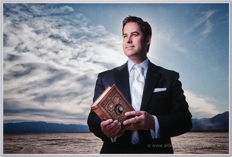 Groom in black suit holds a vintage More Magic book on a dry lake bed beneath moody clouds