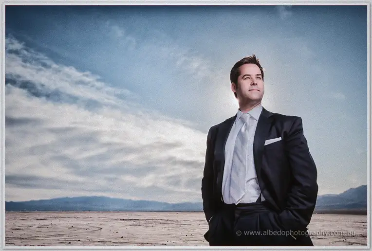 Groom stands hands in pockets on cracked desert flats, bright sun flare behind his shoulders
