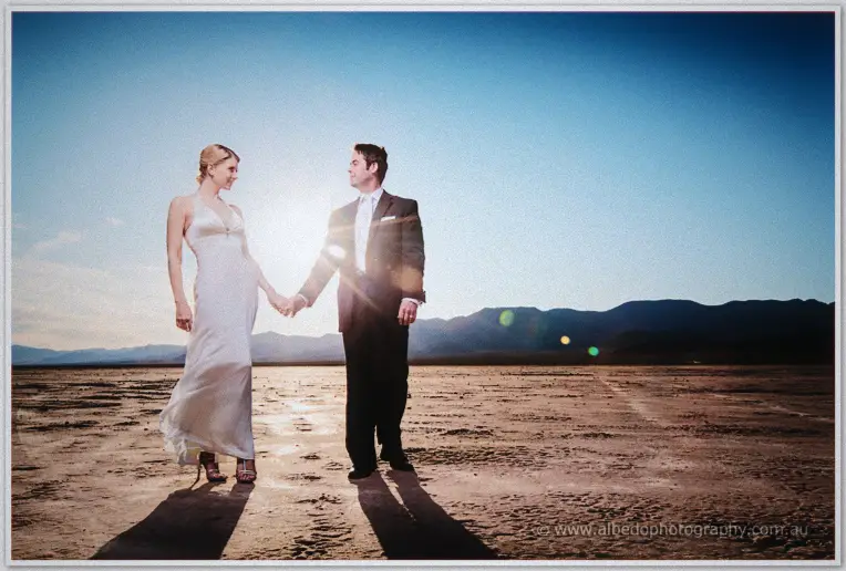 Couple holds hands facing each other, low sun flaring between them on the dry lake bed